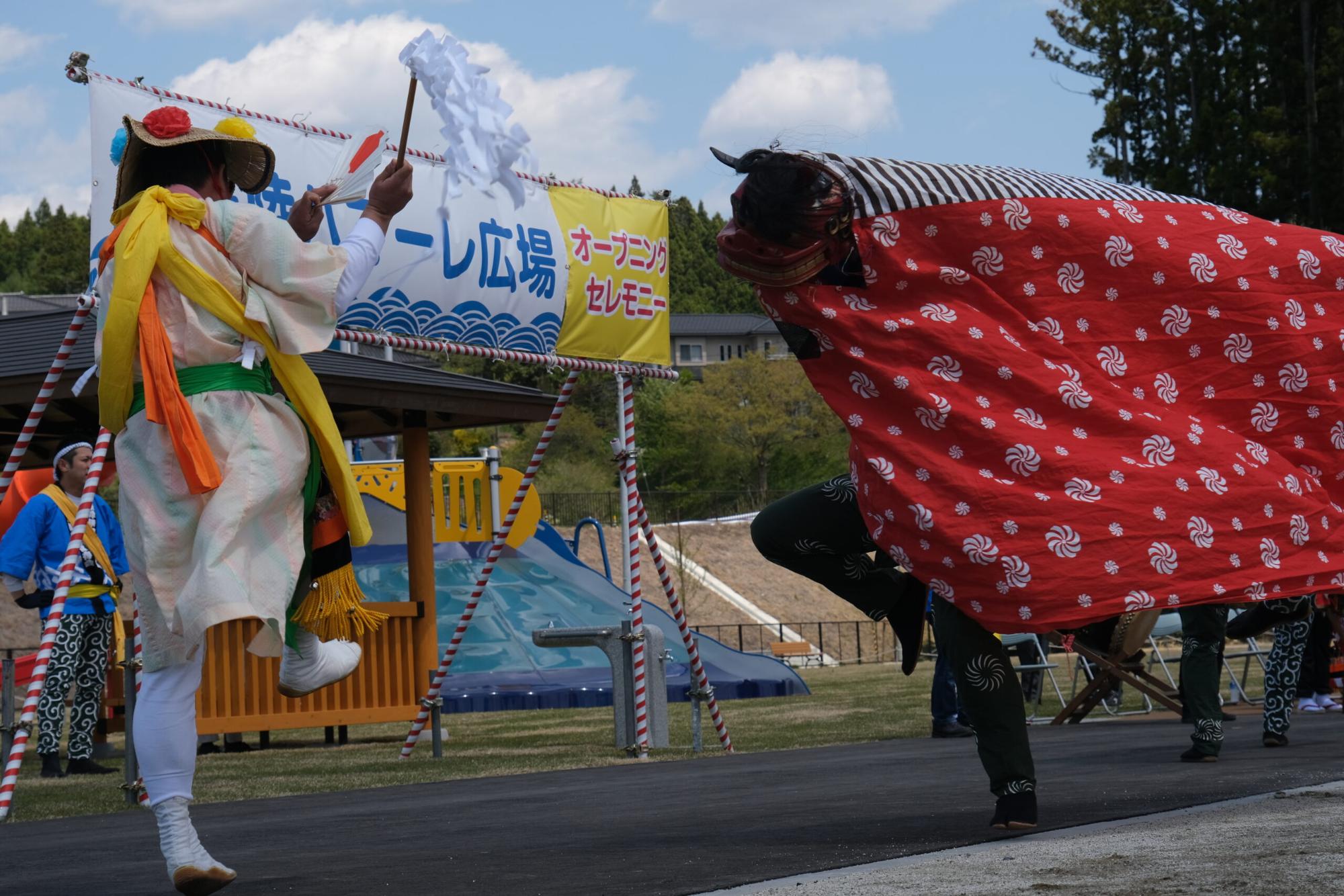 式典の横断幕の前で、伝統的な衣装を着た人物と赤い布で覆われた獅子舞が演技をしている写真