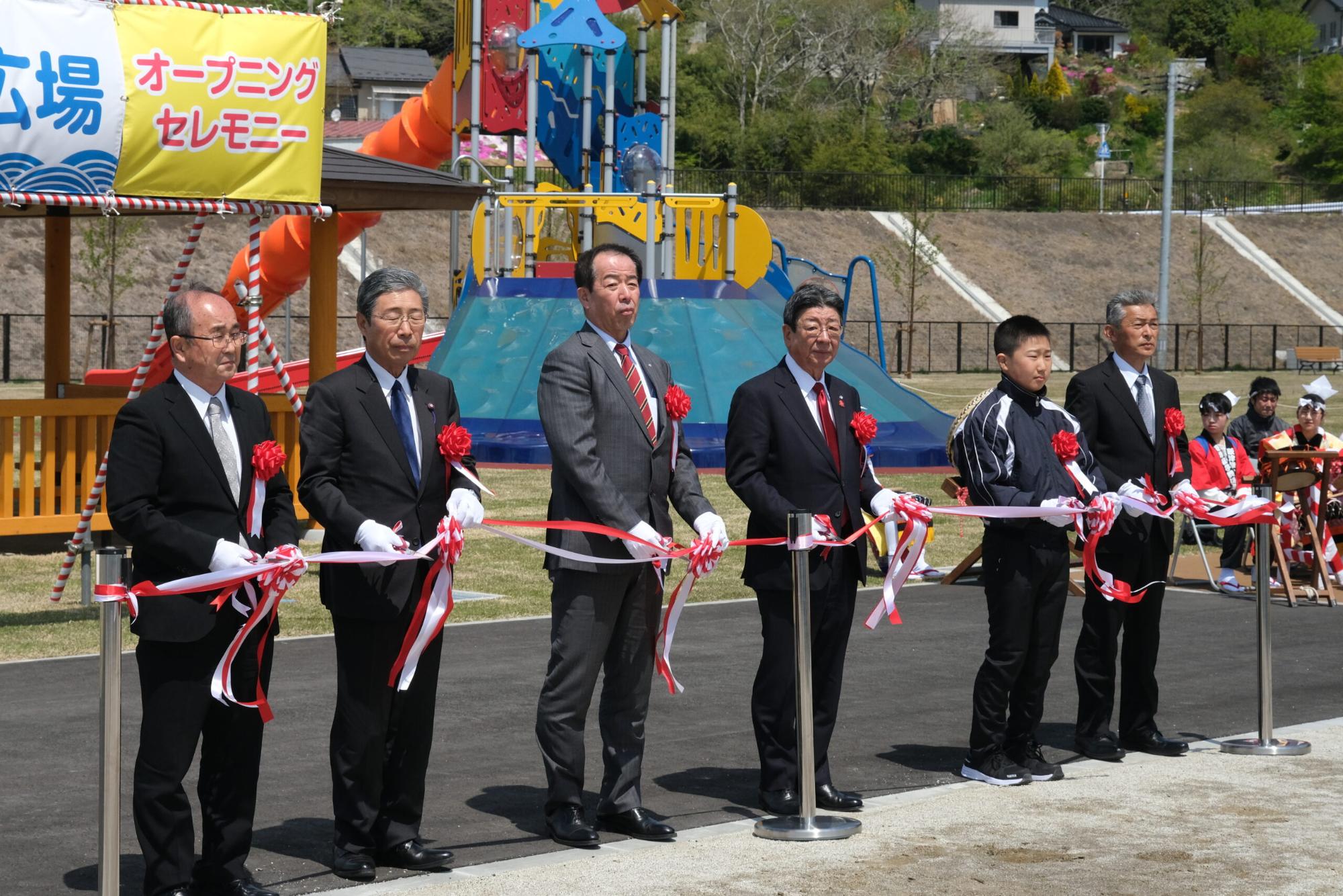 大型遊具がある広場の前で、スーツ姿の男性5人とジャージを着た少年が並び、白い手袋をしてテープカットを行っている写真