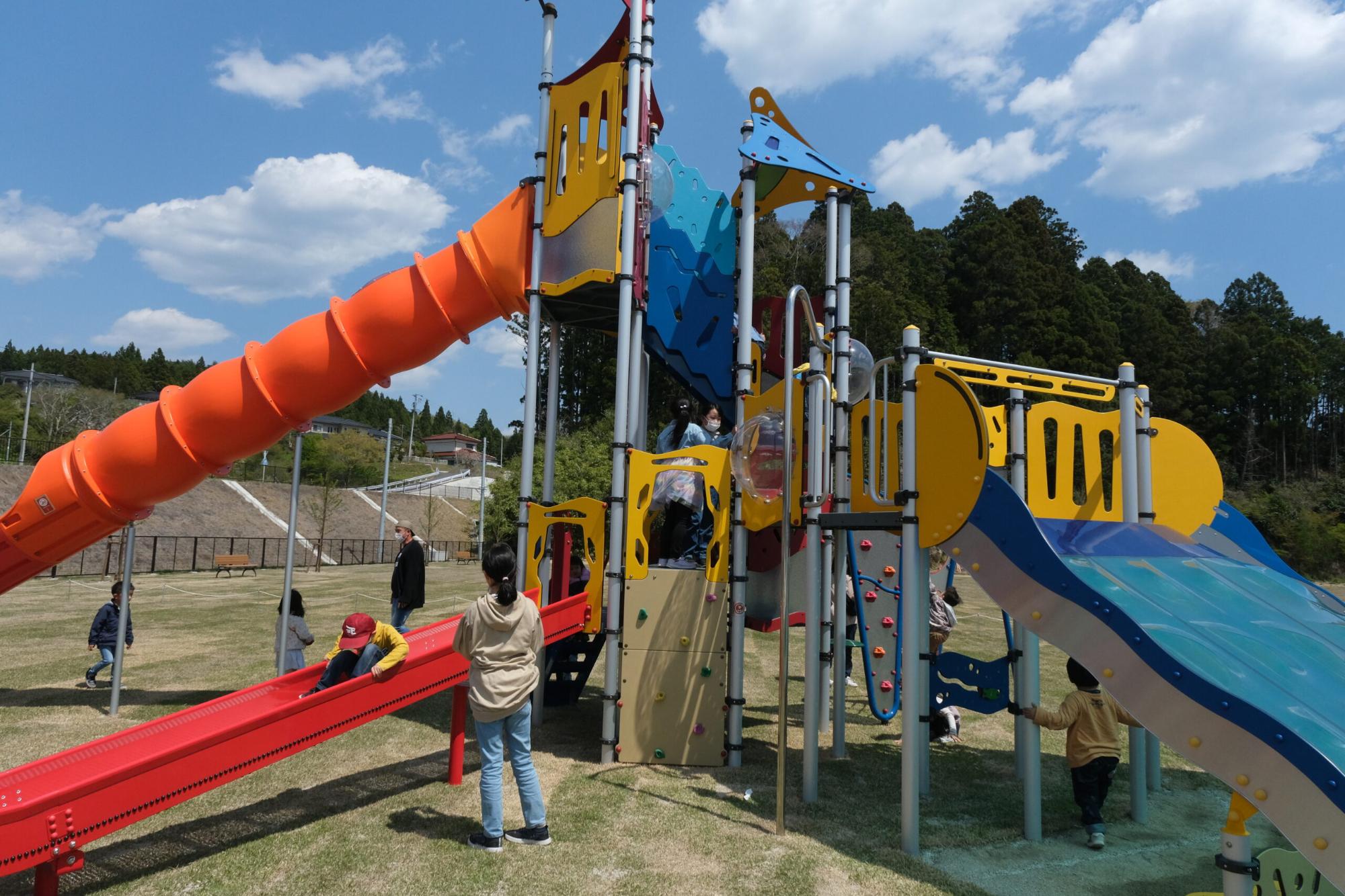 保護者が見守る中、3つの滑り台がついたカラフルな大型遊具で子どもたちが遊んでいる写真