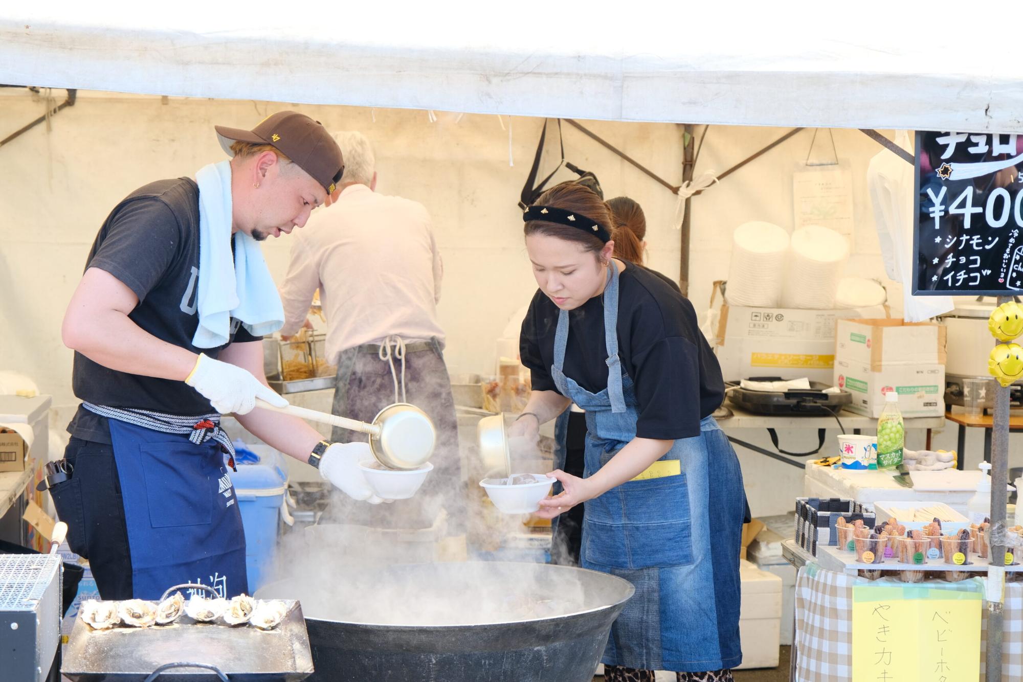 男女2人のスタッフが、テントの下で湯気が上がっている大きな釜から白い容器にスープをよそっている様子の写真