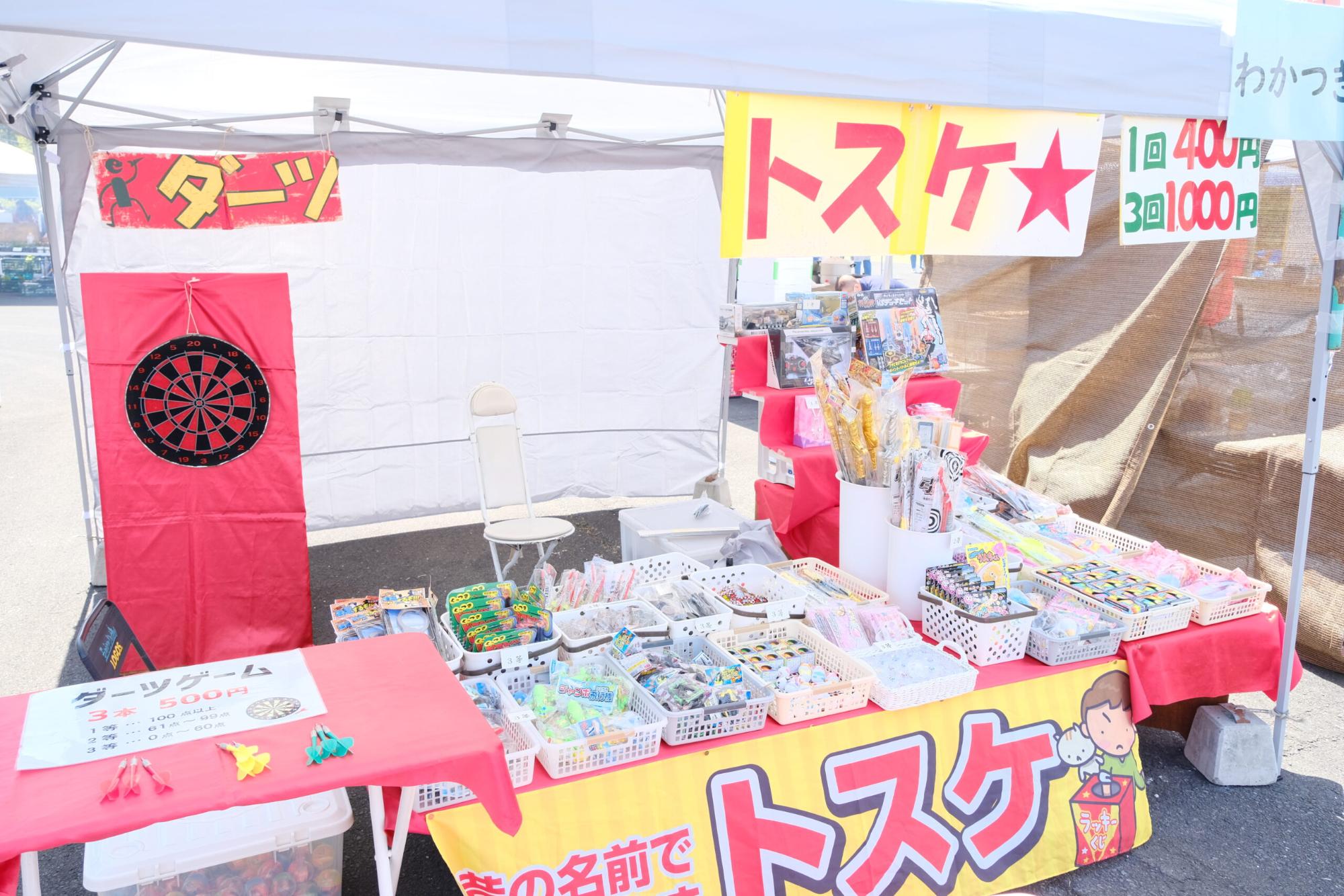 「トスケ」と書かれた看板が設置された、赤いダーツボードがあり、手前の台にはたくさんの景品が並ぶ屋台のテントの写真