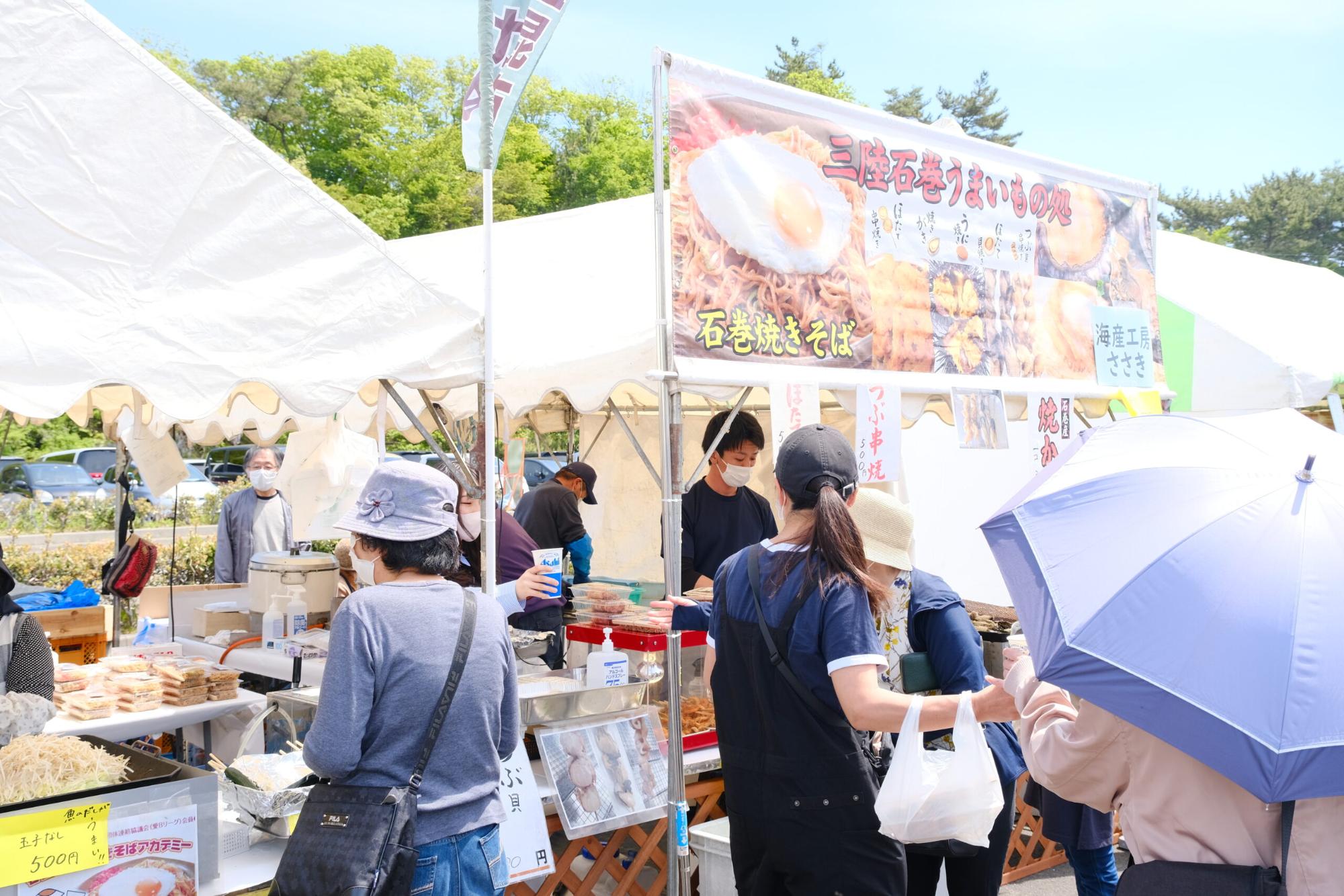 白いテントの前に「石巻焼きそば」などの看板が設置され、たくさんの来場者が料理を求めて集まっている様子の写真
