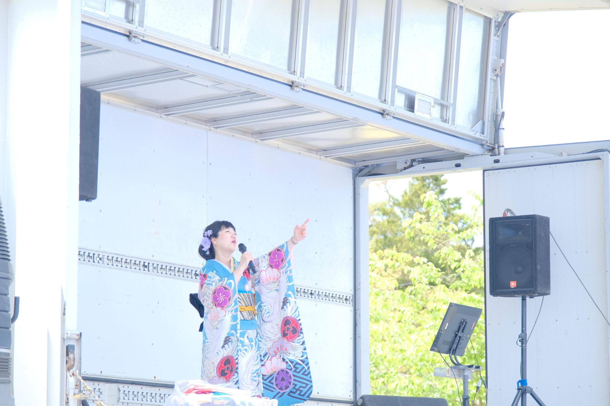 水色を基調とした振袖を着た女性がステージの上でマイクを持ち、左手で空を指さし力強く歌っている様子を撮影した写真