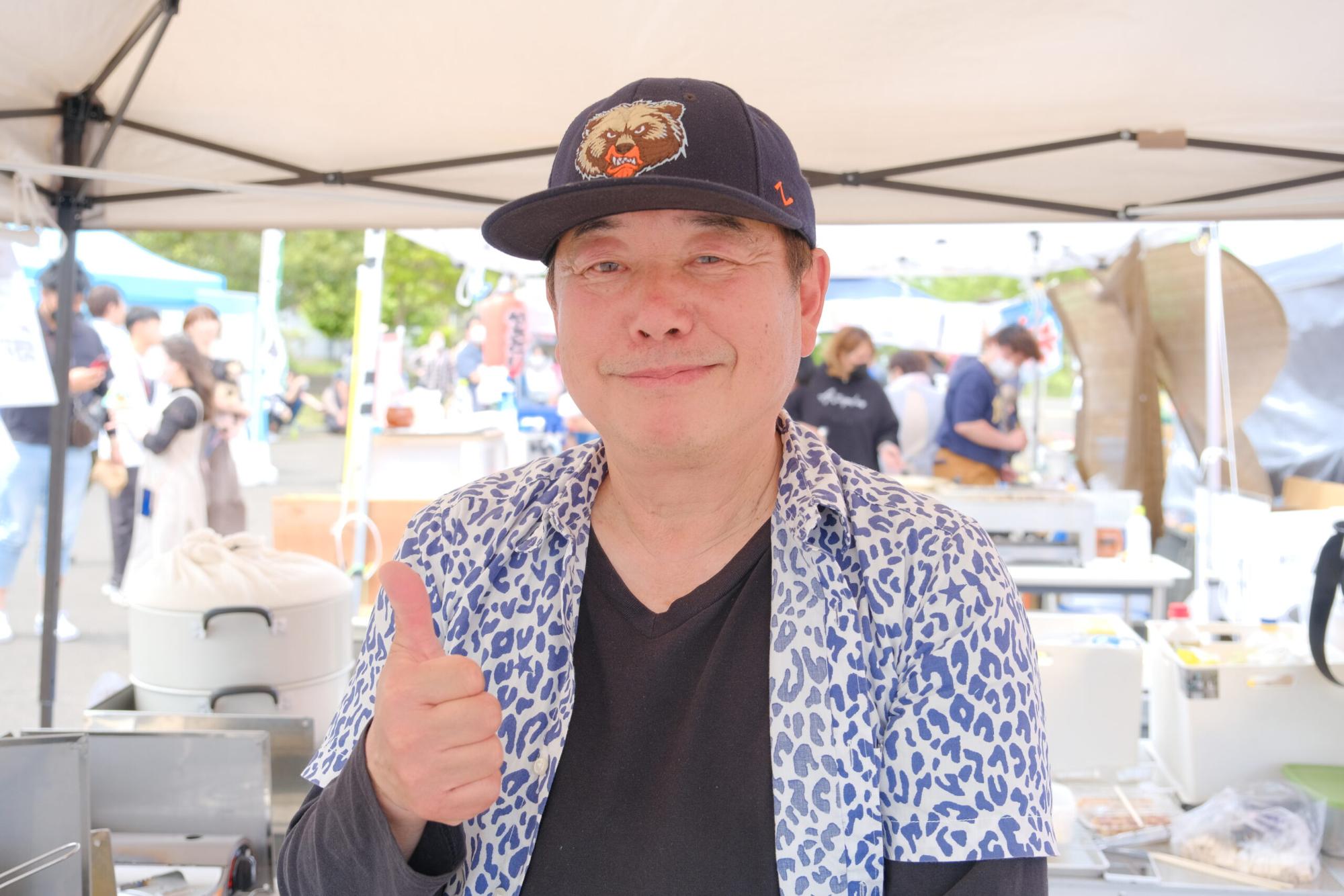 黒い帽子をかぶり青いヒョウ柄のシャツを着た男性が親指を立てて笑顔を見せている写真