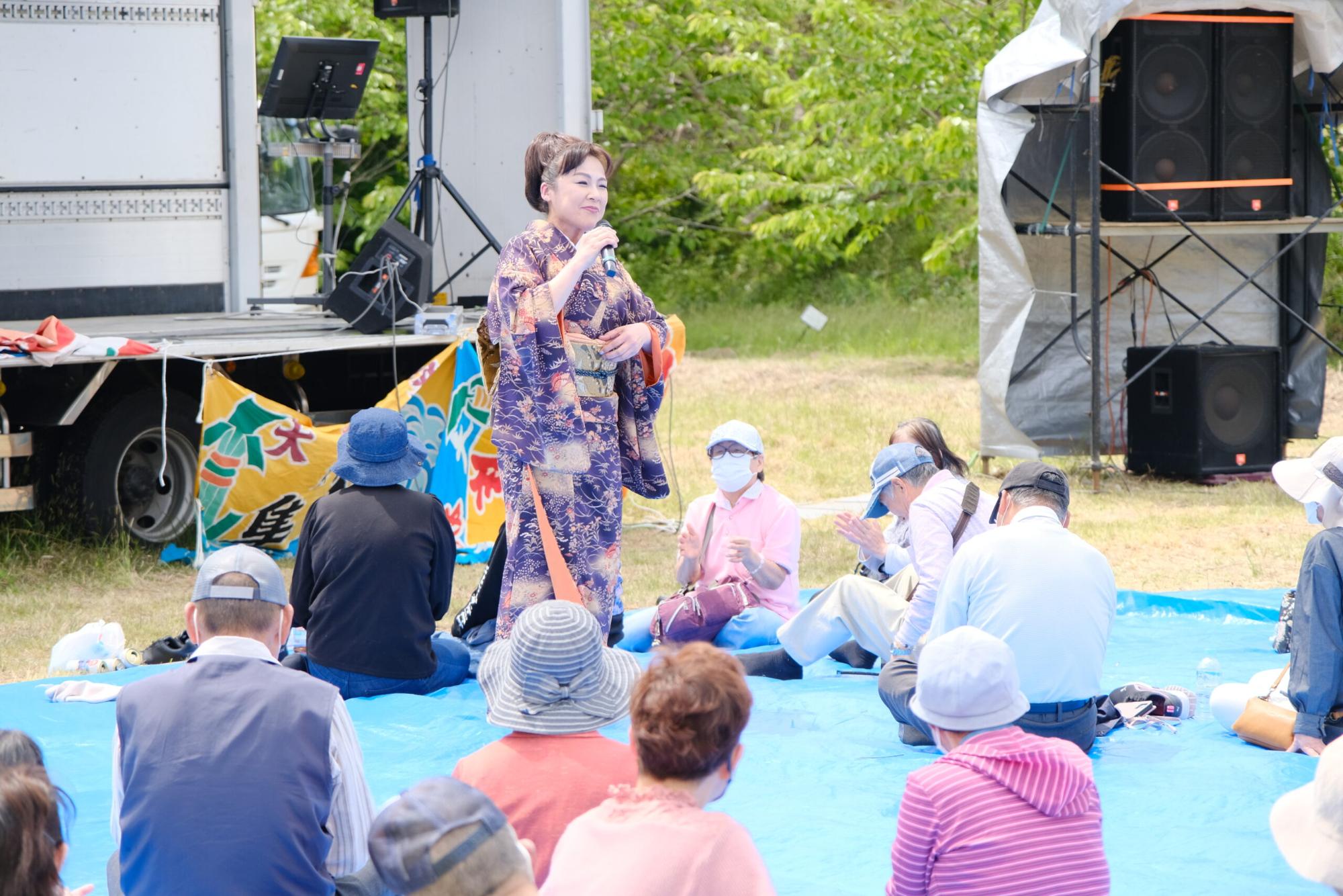 紫色の着物を着た女性がマイクを持って歌を歌っており、レジャーシートの上に座った観客が手拍子をしている写真