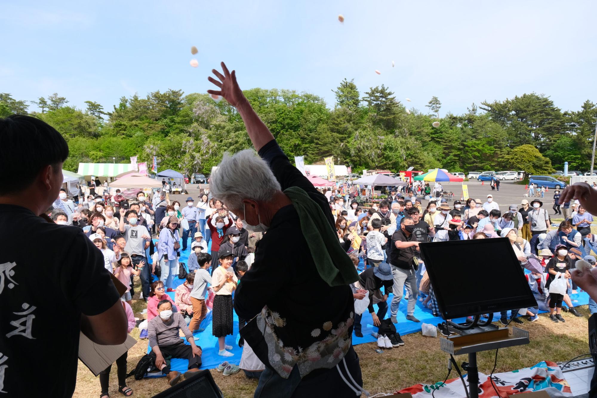白髪の男性がステージの上から手を高く上げて、集まった大勢の来場者に向かって餅やお菓子をまいている様子の写真