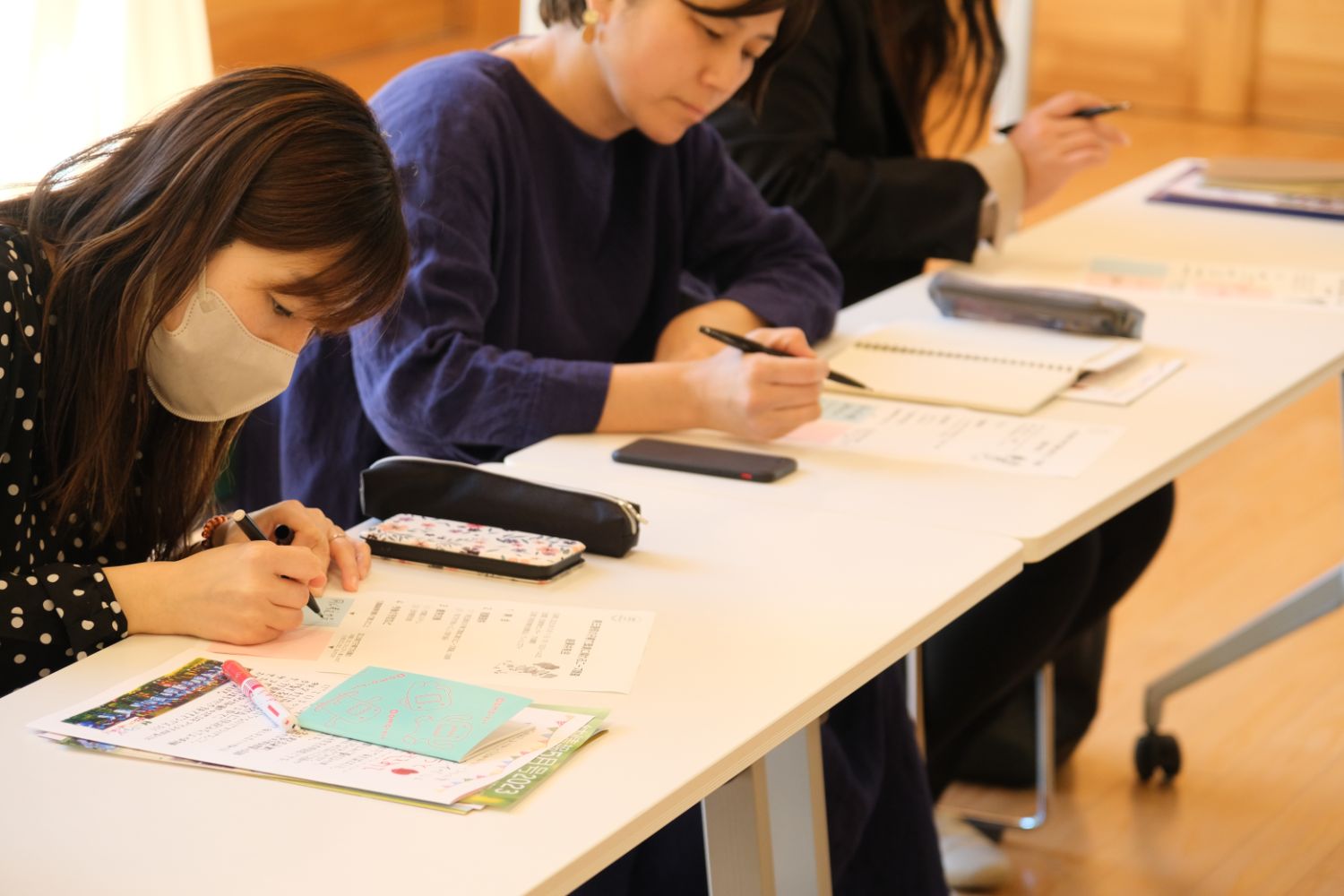 机に座った数人の女性が真剣にペンを動かし、メモや資料に書き込んでいる様子の写真