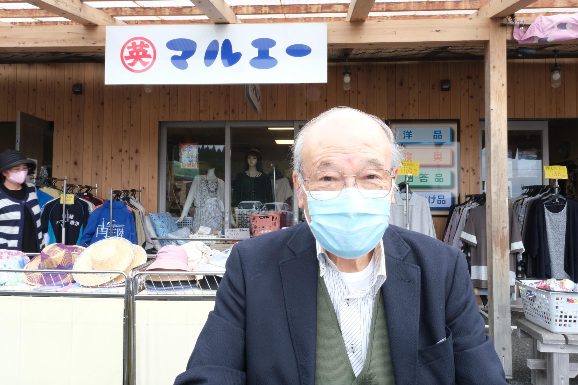 「マルエー」と書かれた店の前で、水色のマスクを付けた千葉委員長が立っている写真