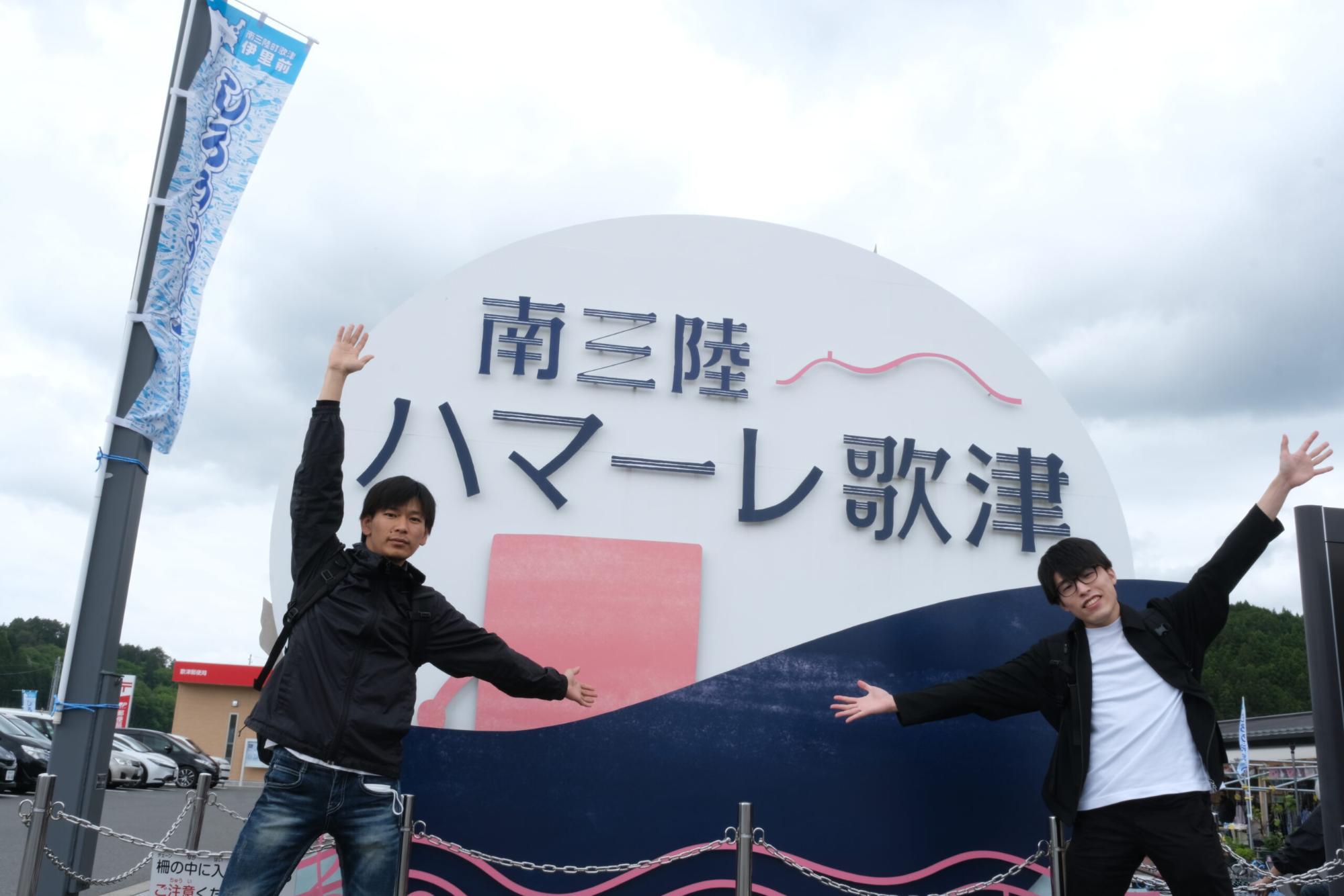 「南三陸ハマーレ歌津」と書かれた丸い看板の前で、2人の男性が両腕を広げて立っている写真