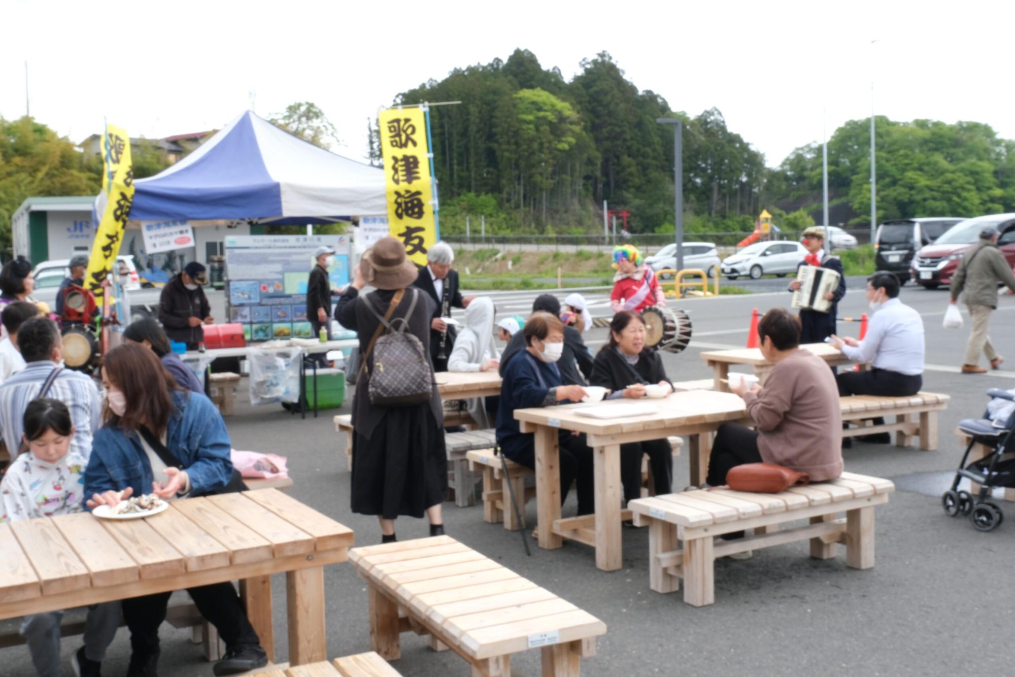 屋外の広場で、複数の木製ベンチとテーブルに人々が座り、食事や会話を楽しんでいる様子の写真