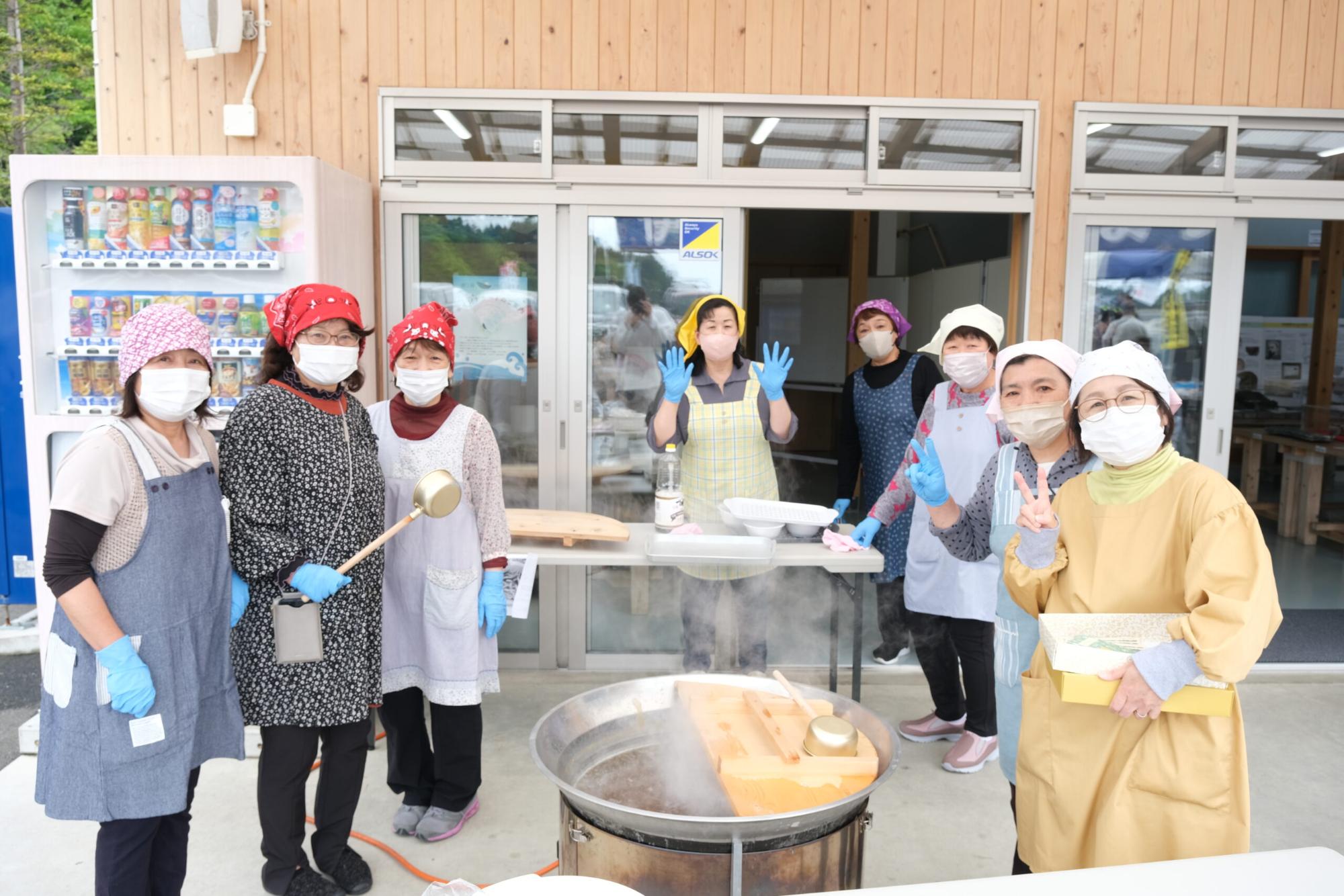 皆エプロンとマスク、手袋を着用した8人の女性が、建物の外に置かれた大きな鍋の周りで、笑顔でカメラに向かってポーズをとっている写真