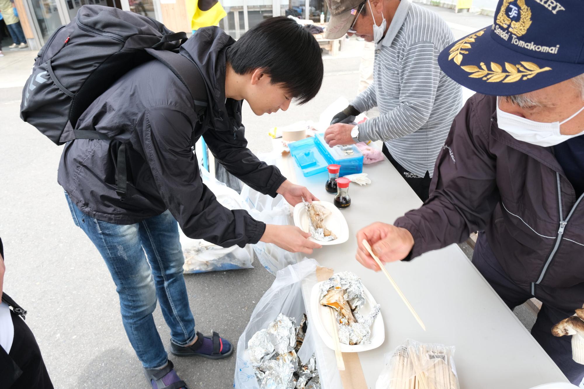 いっちーが屋外のテーブルで焼き魚を受け取り、地域の二人の男性が魚の準備や配膳をしている様子の写真
