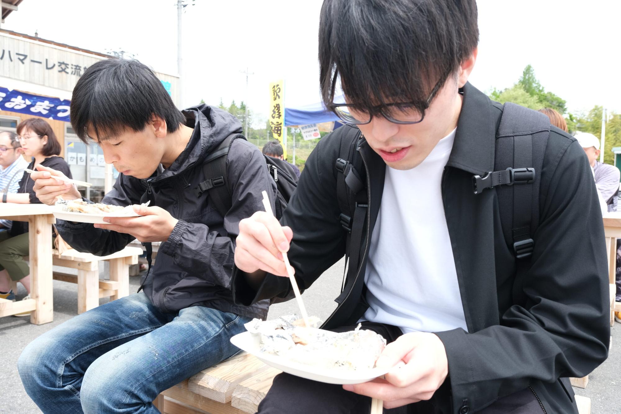 のぶといっちーが屋外のベンチに座って、紙皿に盛られた焼き魚を箸で食べている様子の写真