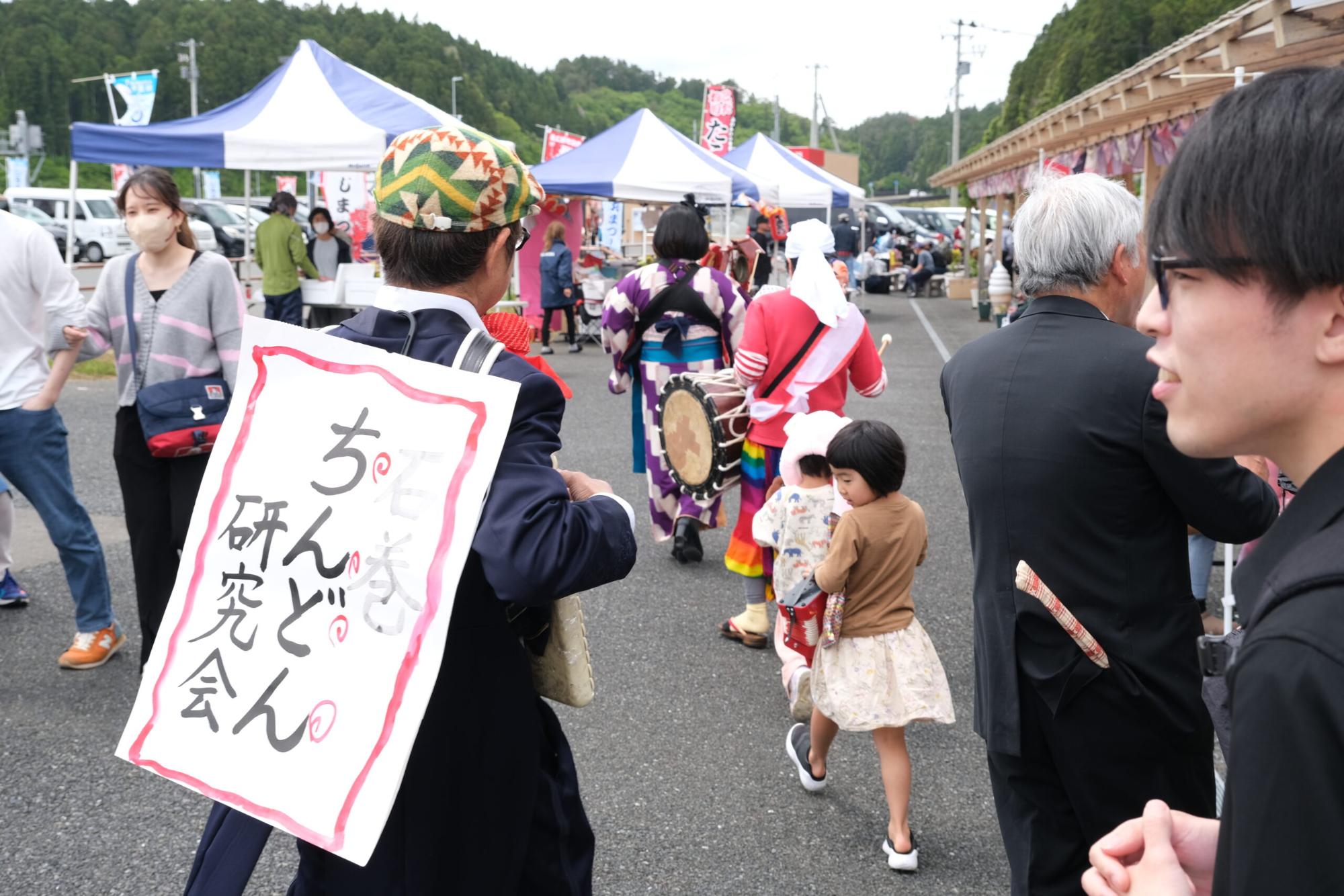 石巻ちんどん研究会の行列や家族連れなどが歩く、屋外のイベントのにぎやかな様子を写した写真