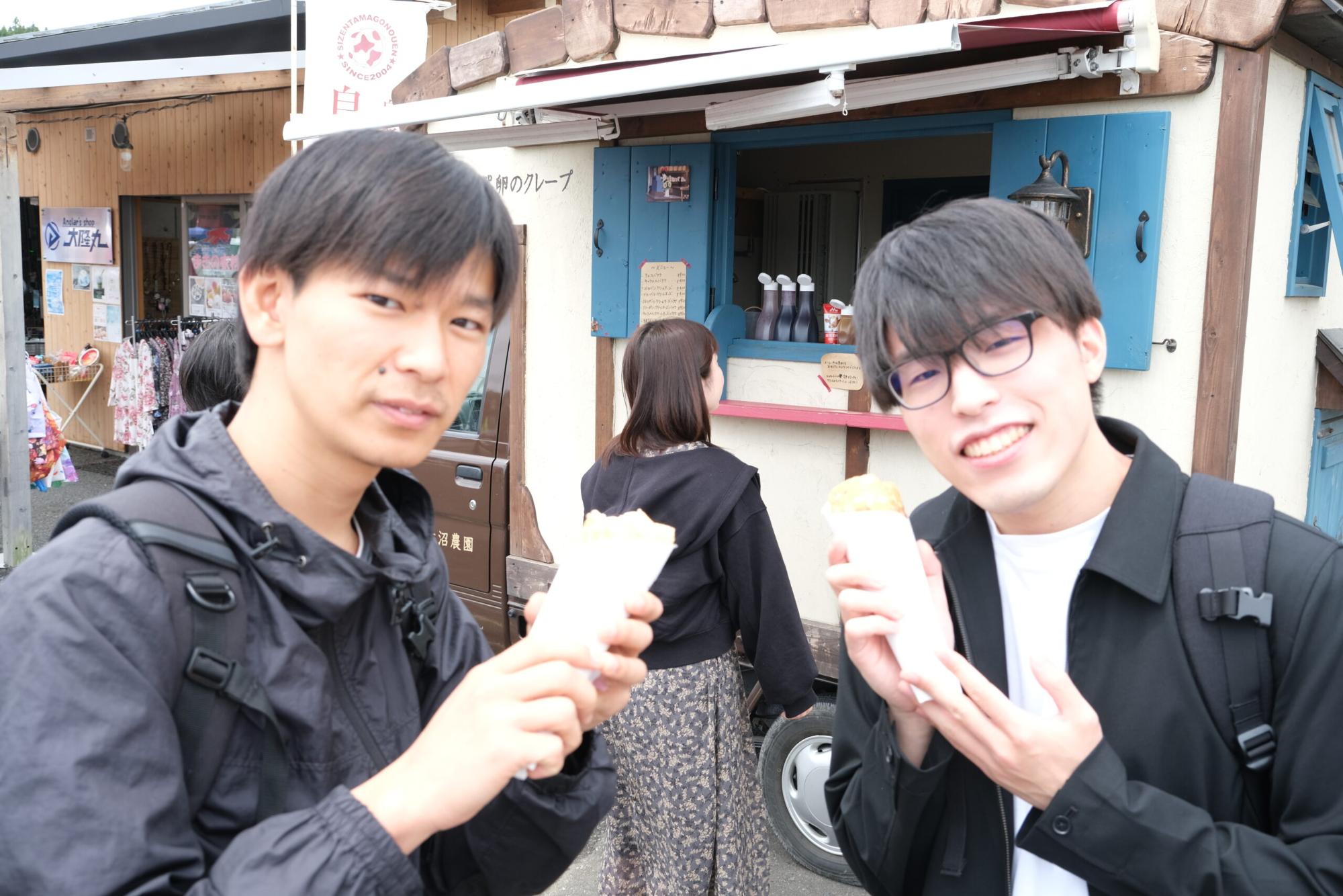 のぶといっちーが屋台の前でクレープを手に持ち、笑顔でカメラに向かっている写真