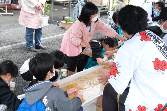 マスクをつけた子どもたちが白い法被を着た男性が座る大きな水槽の周りに集まり、水の中の金魚をすくおうとしている様子の写真