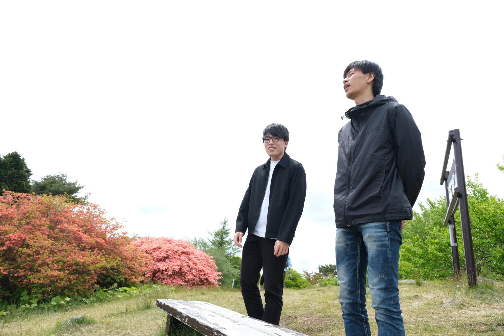 背後にはピンク色の花が咲いたツツジの木々と緑の木々が広がっており、高台のベンチの前でのぶといっちーが立っている写真