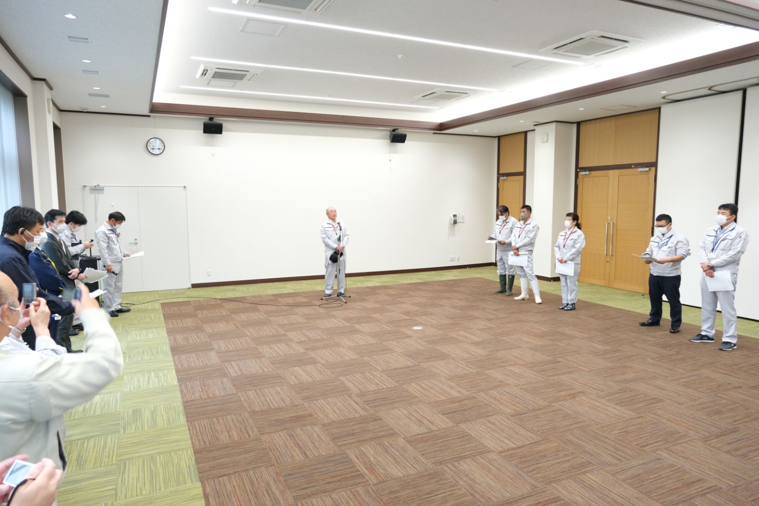 室内の広い会議室で、作業服を着た複数の人々が左右に分かれて立ち、中央で男性がマイクの前に立って話している写真