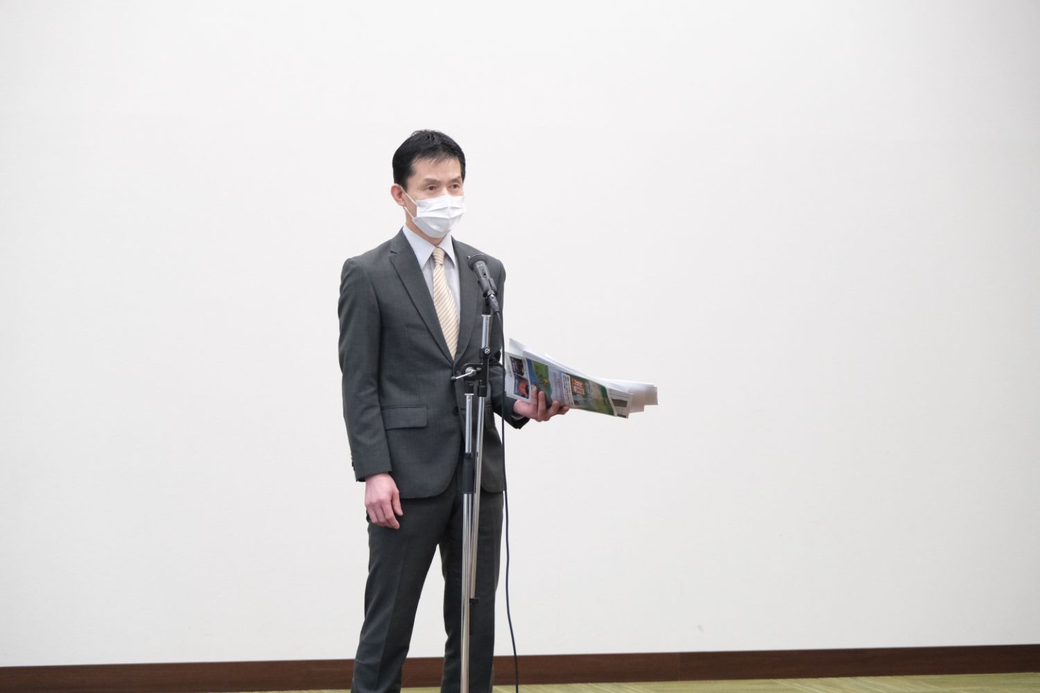 スーツ姿の男性がマイクの前に立ち、資料を持ちながら話している写真