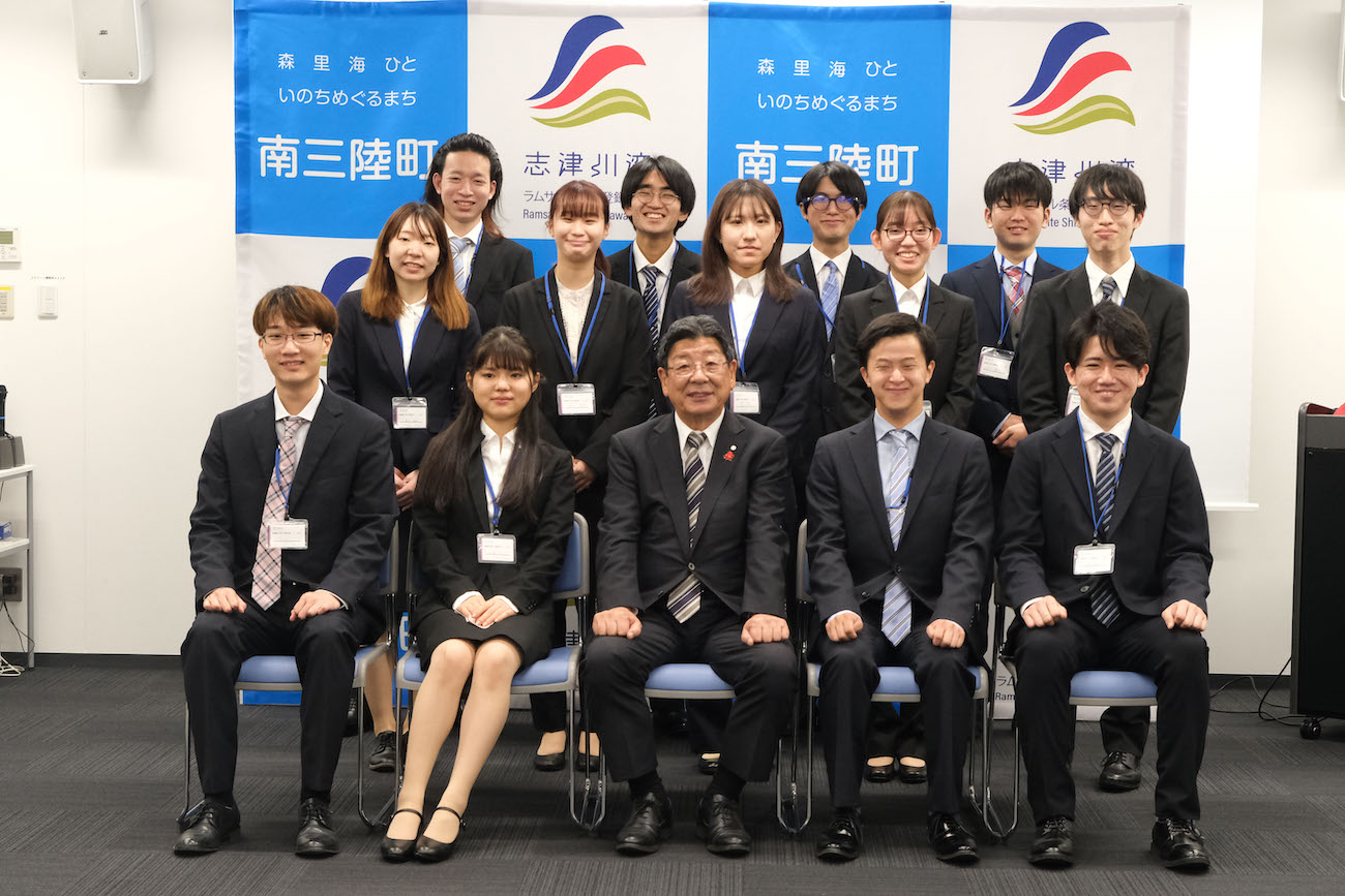 バックパネルの前でスーツ姿の大学生と町長と関係者が並んだ集合写真