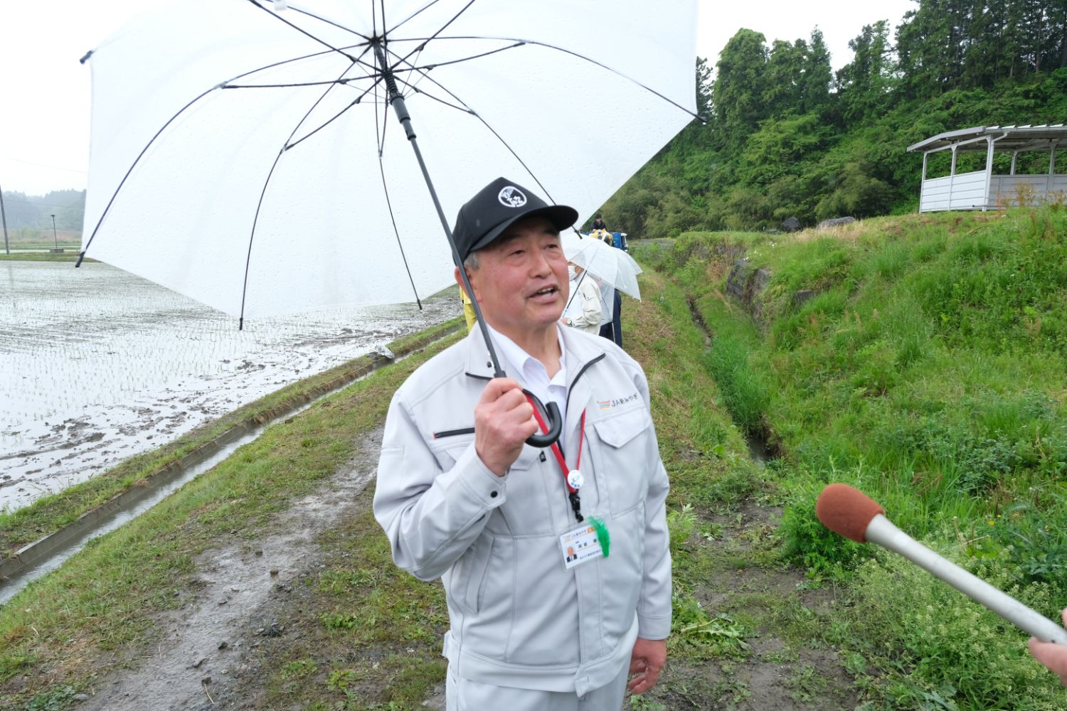 白い傘をさした作業服の男性が田んぼのそばで話をしている写真