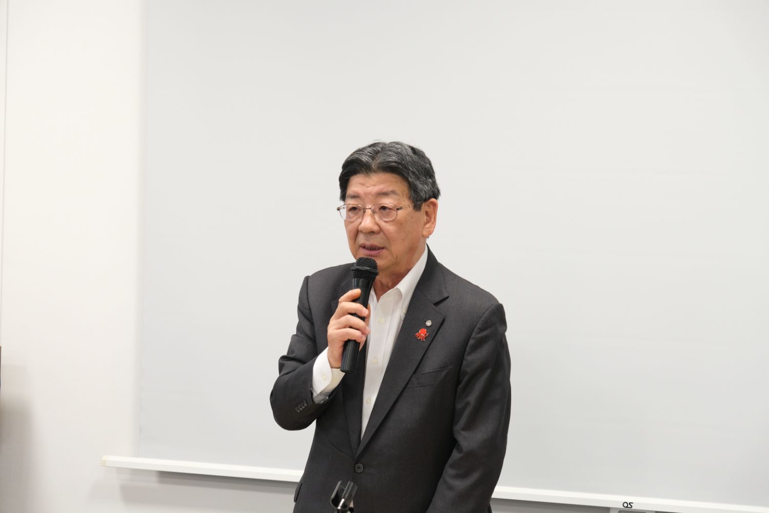 スーツを着た塾長の男性が前に立ち、マイクを持って南三陸町の取り組みについて語っている様子の写真