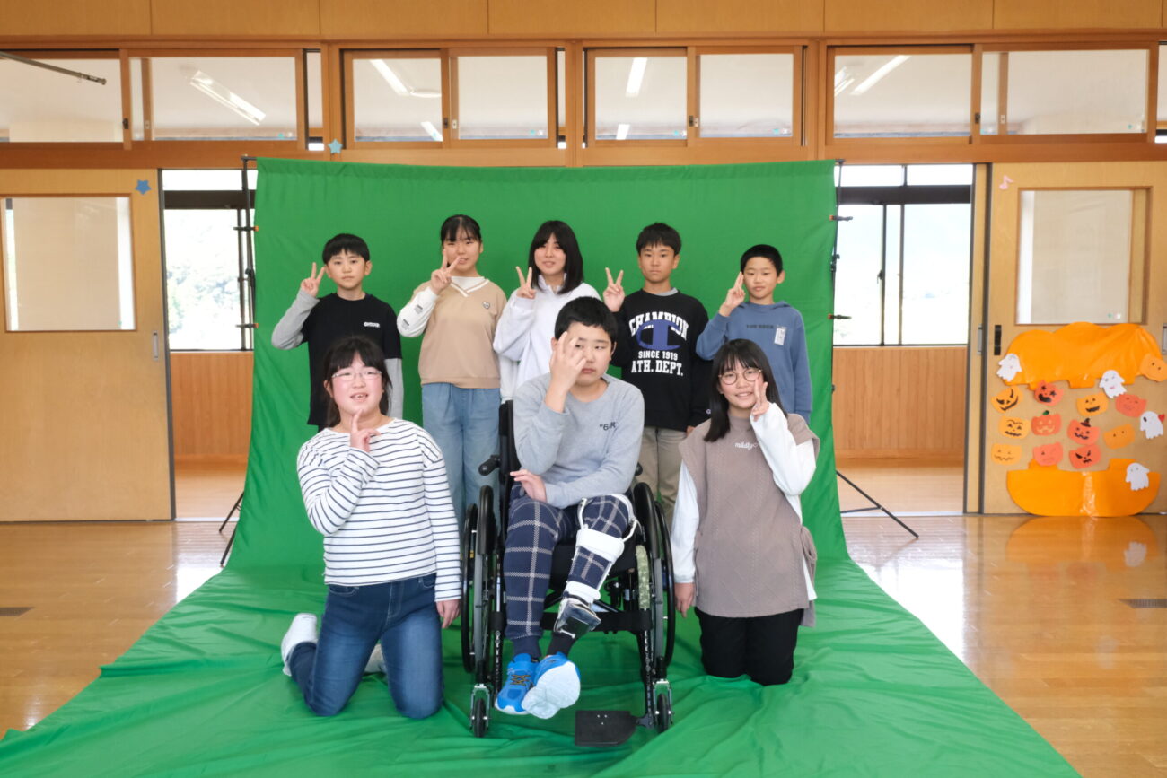 教室に設置したグリーンバックで車椅子の児童を含み子どもたちが笑顔でポーズをとっている写真