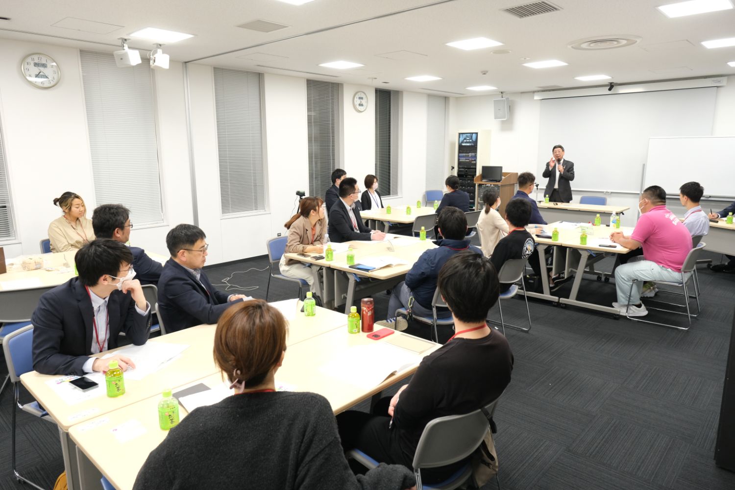 スーツやカジュアルな服装の参加者たちが5つのテーブルに分かれて座り、正面の講師の方を向いて話を聞いている講義の全体風景の写真