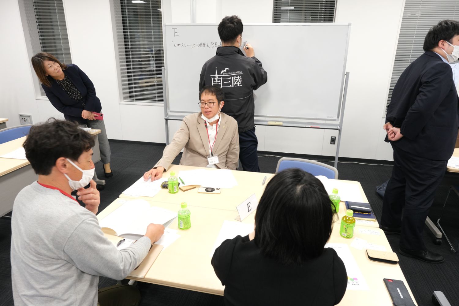 「南三陸」と書かれた上着を着た参加者がホワイトボードに意見を書いており、他のメンバーがテーブルを囲んで話し合っている様子の写真