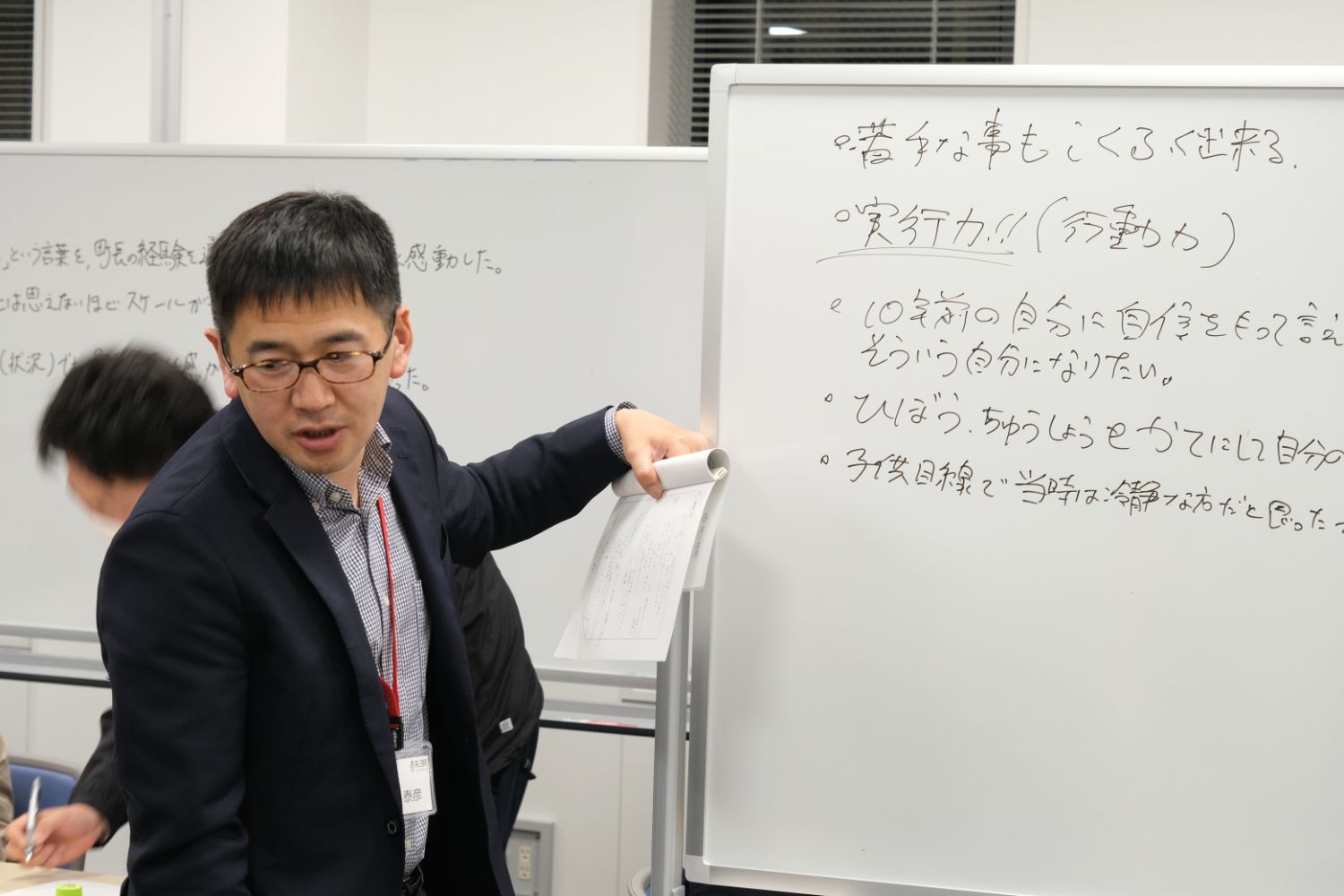 スーツ姿の男性が「実行力」などの言葉が書かれたホワイトボードを指し示しながら、話をしている様子の写真