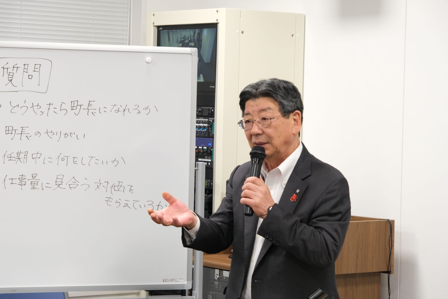 塾長が「町長のやりがい」や「任期中に何をしたいか」などの質問が書かれたホワイトボードの前で、マイクを使って回答を行っている写真