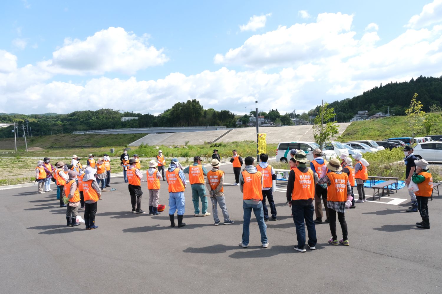 オレンジ色のベストを着た人々が広い駐車場で輪になり、佐藤町長の話を聞いている様子の写真