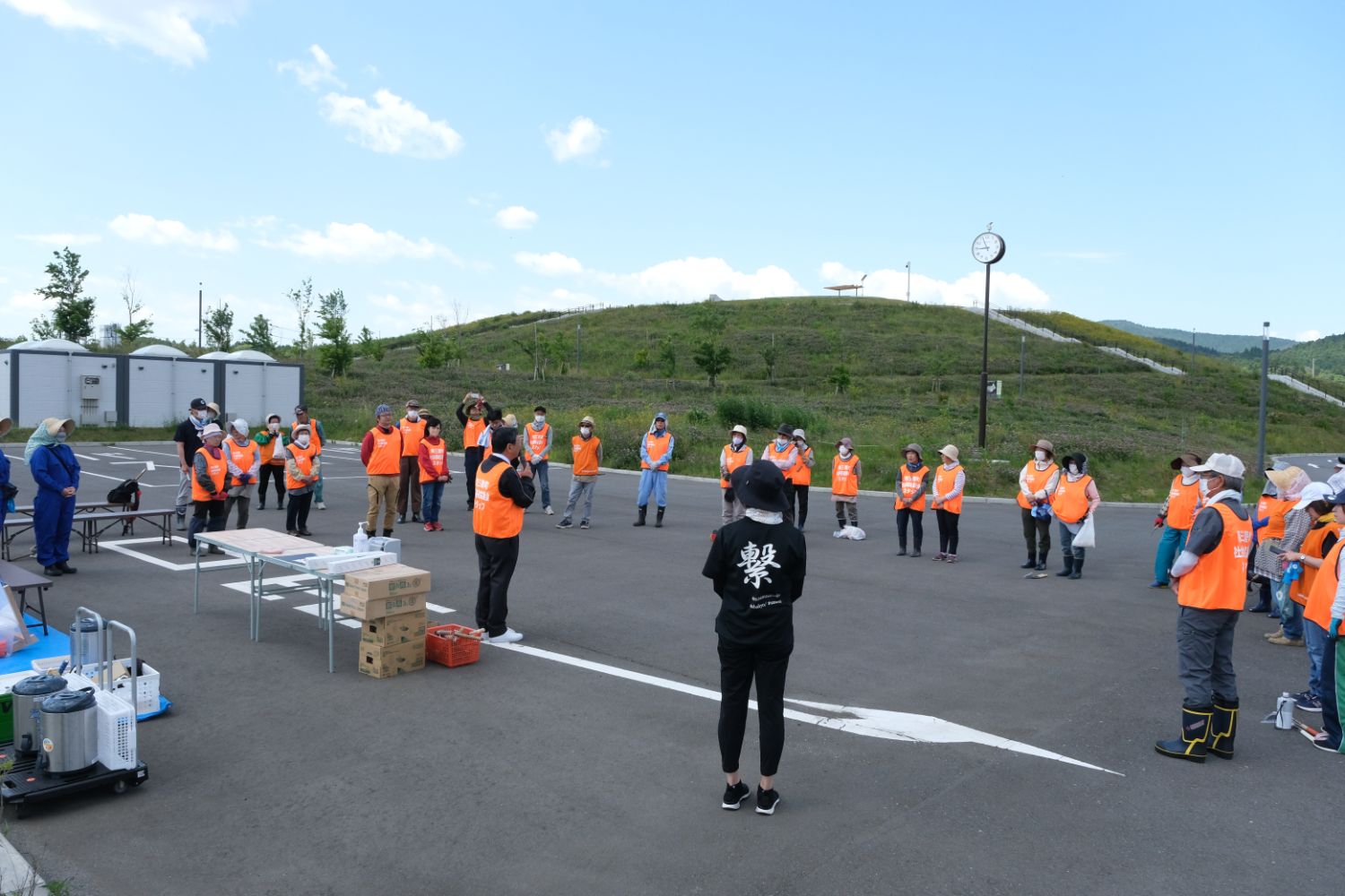 青空の下、オレンジ色のベストを着た人たちが広い駐車場で円を作り、男性が話をしているの方を向いている人々の中に、背中に「繋」と書かれた黒いTシャツを着た人が見える写真