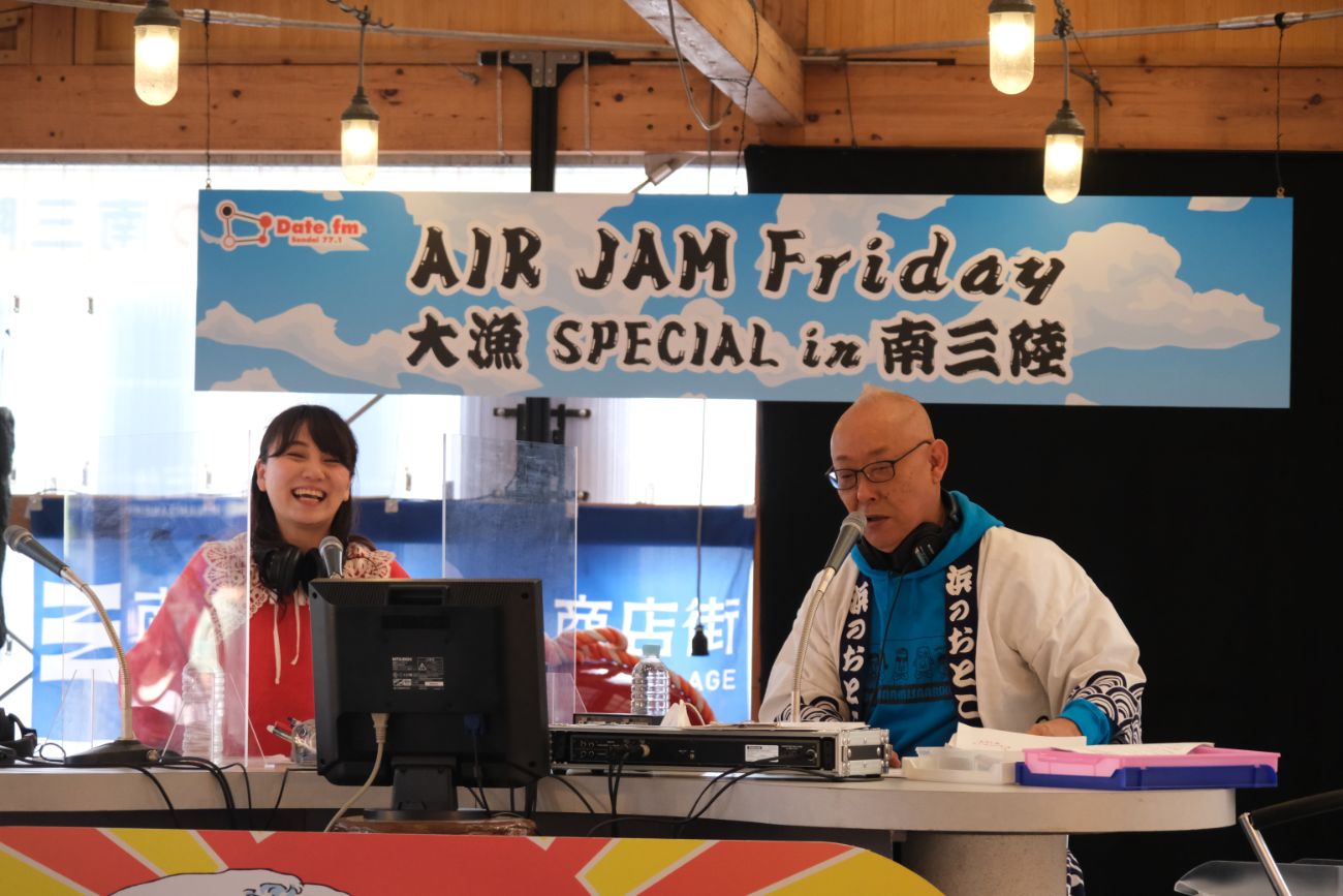「AIR JAM Friday 大漁SPECIAL in 南三陸」のラジオブースで、佐藤友紀乃さんが笑顔で話し、隣の本間秋彦さんがマイクに向かって真剣に話している様子がアップで写されており、番組の進行中の活気が伝わる写真