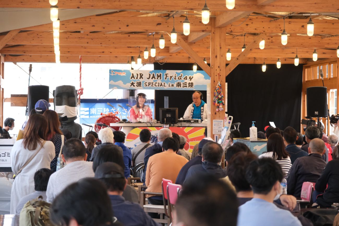 多くの観客が集まり、木造の屋根の下で「AIR JAM Friday 大漁SPECIAL in 南三陸」の公開ラジオ収録を見守っており、舞台にはMCとアシスタントが座って番組を進行している様子が写っている写真