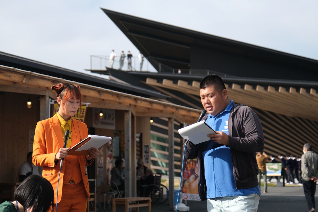 屋外の商業施設前で、オレンジ色のスーツを着たドル萌江さんと青いポロシャツを着た佐藤潤也さんが、それぞれマイクと資料を持ってイベントの進行をしている様子の写真