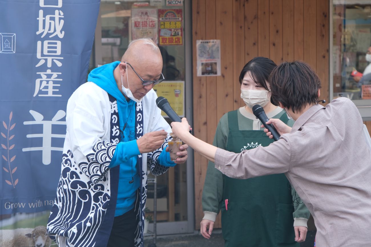 内木渡さんがカップの中の食べ物を見つめており、周囲にはインタビュー用のマイクを持った男性と、エプロン姿の女性が立っているインタビューの写真