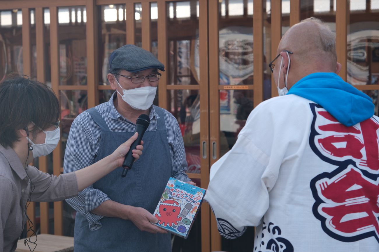 屋外で「AIR JAM Friday 大漁SPECIAL in 南三陸」のMCが、緑のエプロンを着た地元の男性とインタビューをしており、MCが手に持った「オクトパス君せんべい」を見ながら話す様子の写真