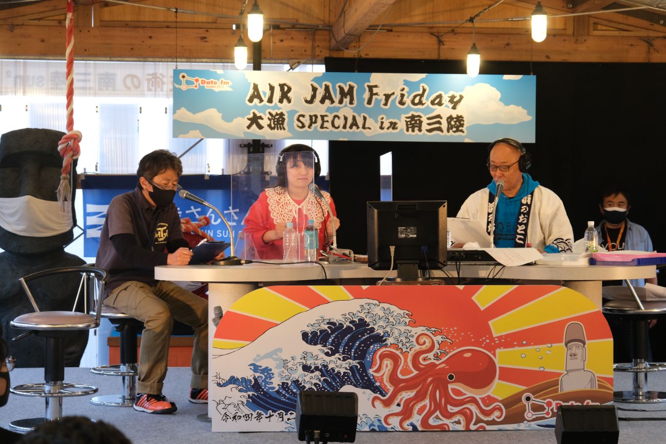 「AIR JAM Friday 大漁SPECIAL in 南三陸」の公開収録の舞台で、MCとアシスタントの女性、ゲストの内木渡さんがマイクの前に座って番組を進めている様子の写真