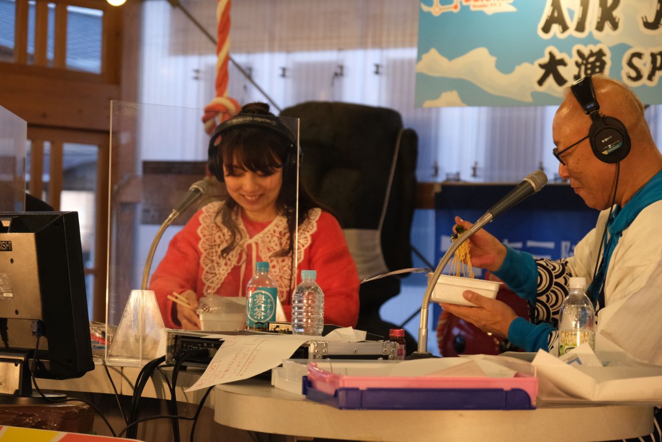 ラジオ番組中、男女2人が透明なパネル越しに向かい合い、食事をしながら楽しそうに話している写真