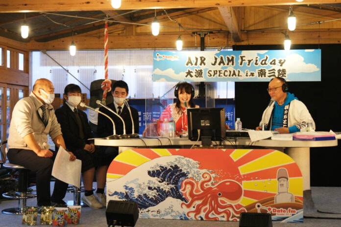 地元の学生や大人たちがマイクの前に座り、「AIR JAM Friday 大漁SPECIAL in 南三陸」の特設ラジオブースでインタビューを受けており、背景には手作りの看板や太陽と波、タコなどが描かれたパネルがあり、地域と漁業をテーマにしたラジオ番組が公開収録されている様子の写真