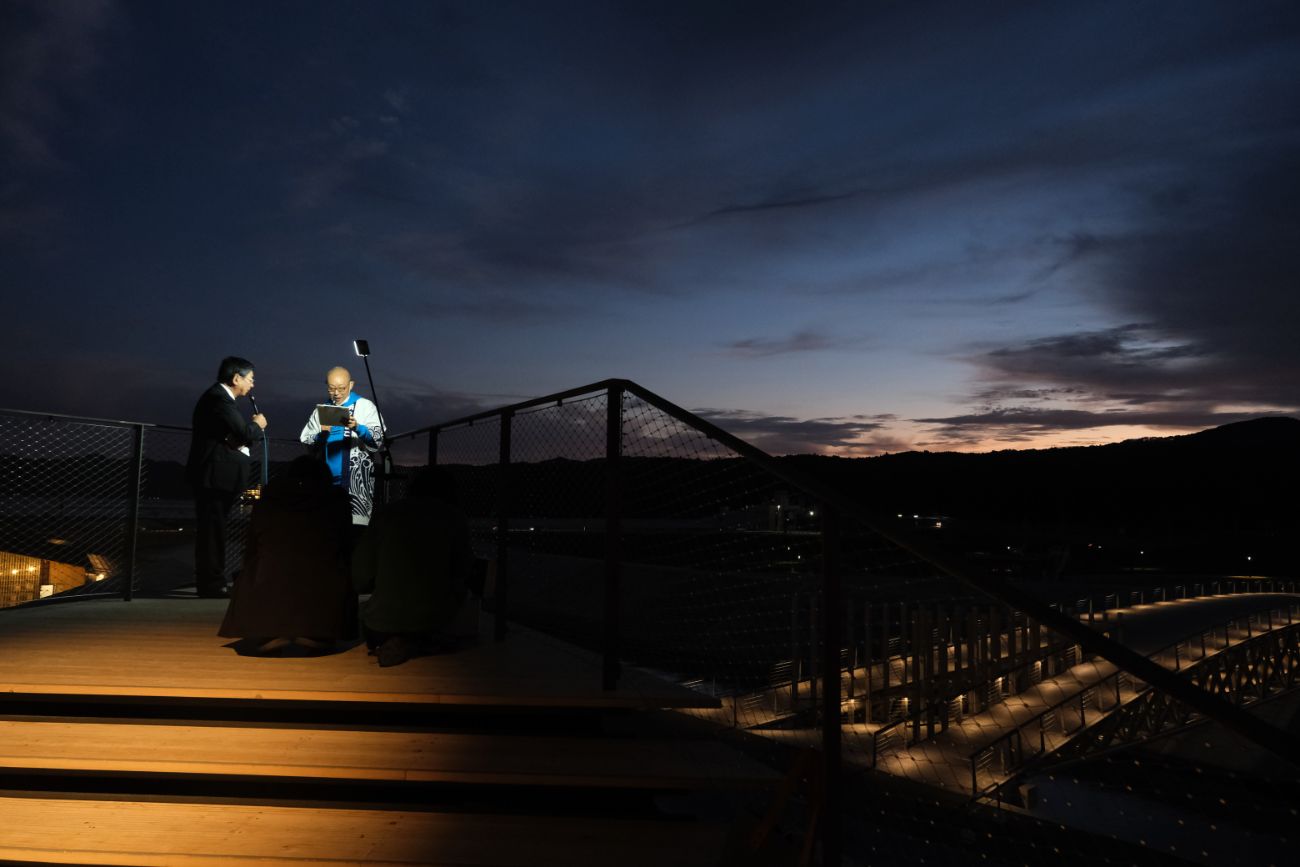 夜の屋外ステージで、照明に照らされた2人の男性がマイクを使って話している写真