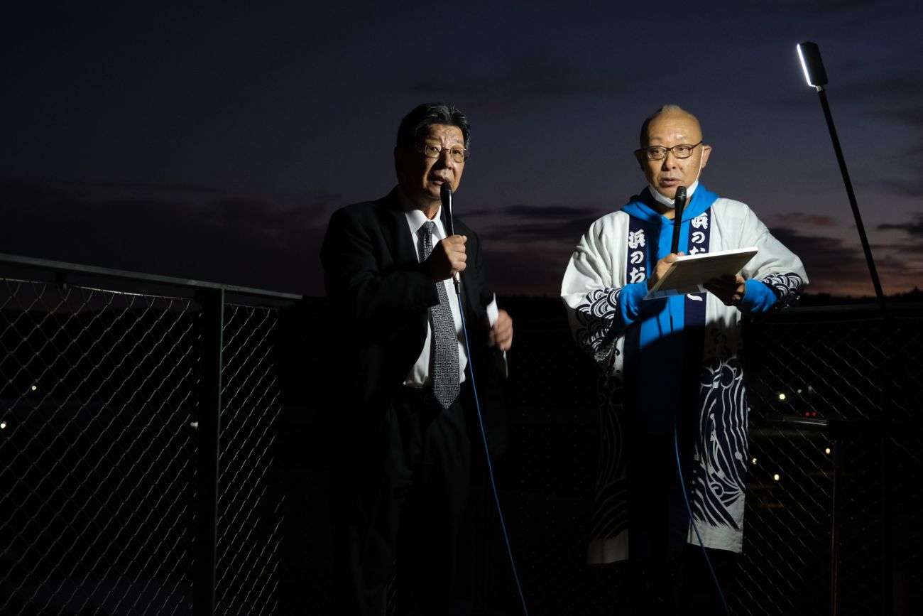 スーツ姿の佐藤仁町長と法被を着た内木渡さんが、夜景を背景に並んでインタビュー中の様子の写真