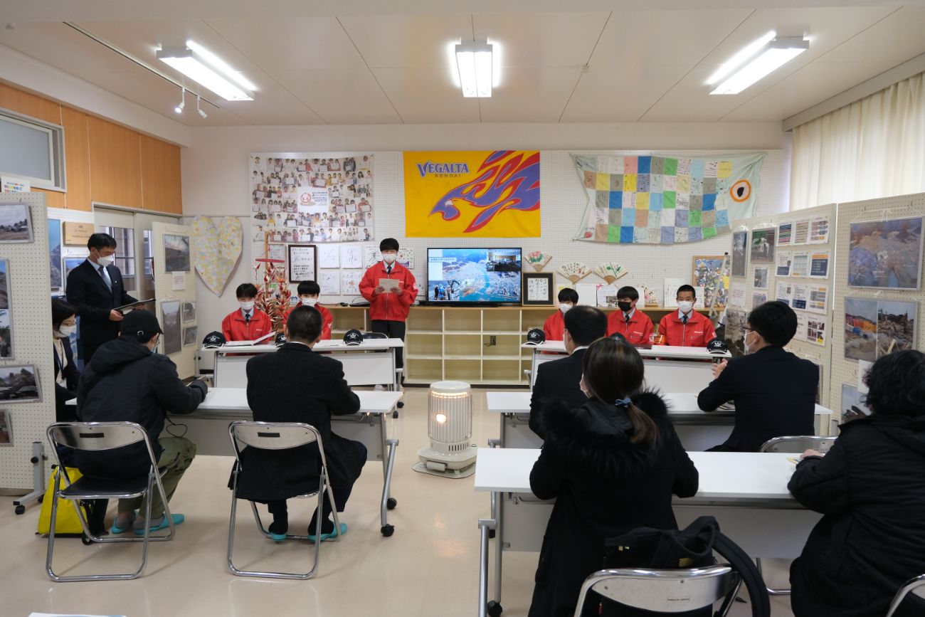 前方に並んだ赤いジャンパー姿の生徒たちが発表を行い、それを聞く関係者の方々が長机の席に向かい合って座り、意見交換をしている様子の写真