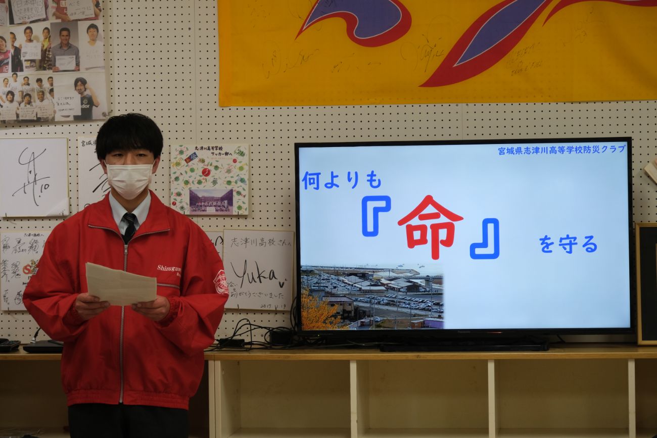 赤いジャンパー姿の男子生徒が資料を持ち、背後のモニターに「何よりも命を守る」と映る発表中の写真