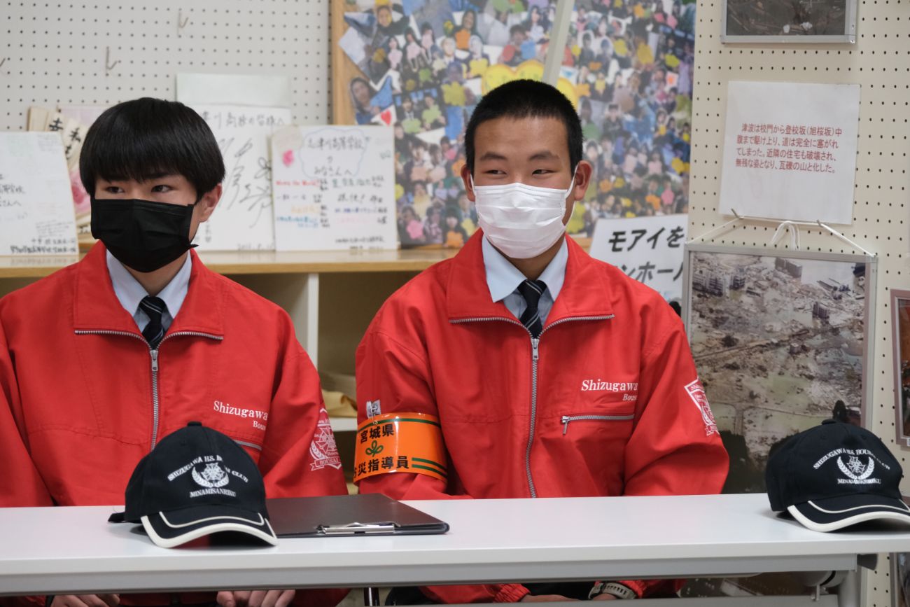 赤いジャンパー姿の男子生徒2人が並んで座り、前の机に黒い帽子が置かれている様子の写真