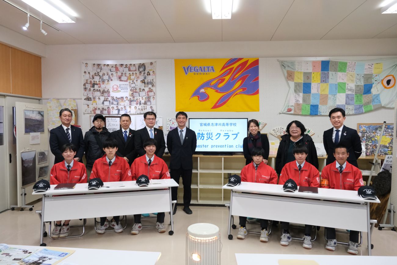 赤いジャンパー姿の生徒6人が机に座り、その後ろにスーツ姿の関係者の方々が立つ集合写真