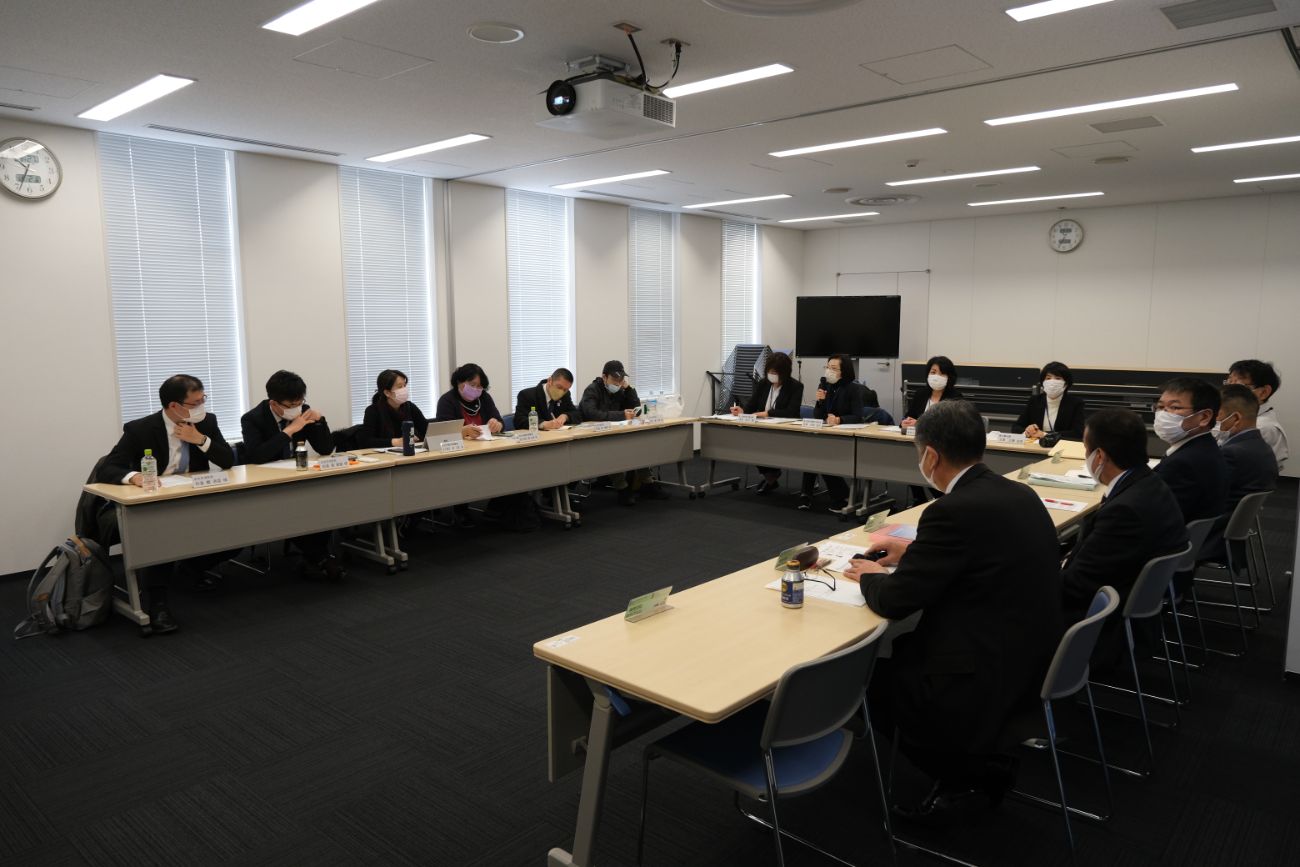 長机をコの字に並べた会議室で、スーツ姿の関係者の方々がマスクを着用し資料を見ながら会議をしている様子の写真