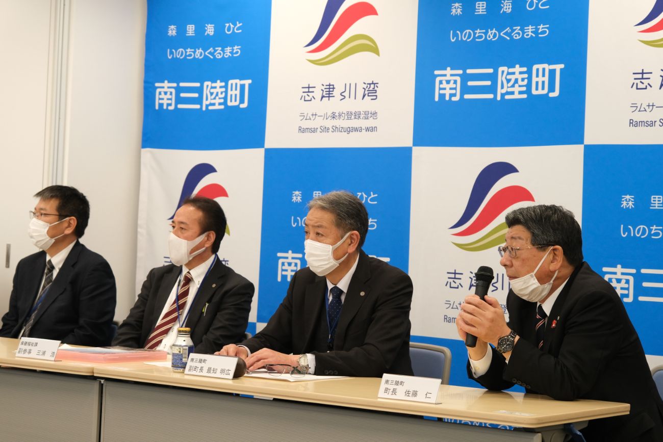 南三陸町や志津川湾のロゴが描かれた背景前で、スーツ姿の男性4人がマスクを着用して並び、町長がマイクを持って発言している様子の写真