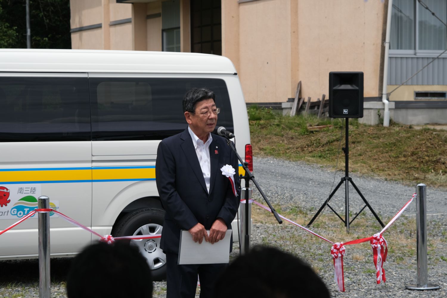 赤白のテープで仕切られた式典用のスペースに停まっている白いワゴン車の前で、スーツ姿の男性が屋外でマイクを使って話している写真